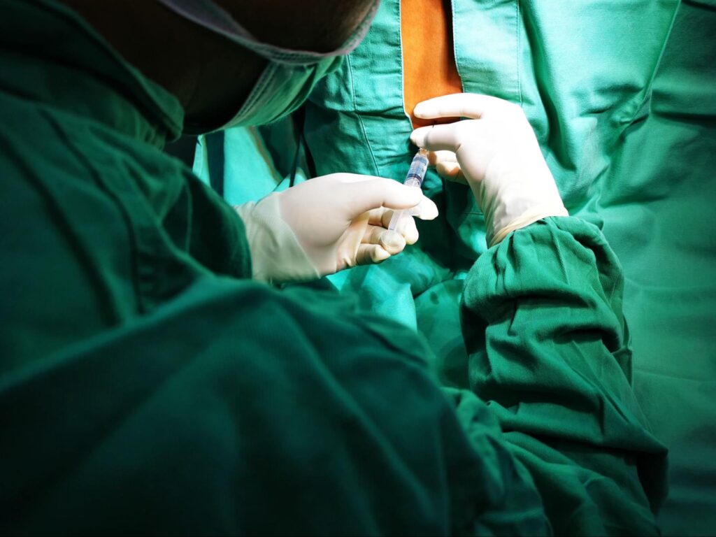 A CRNA in green scrubs injects an epidural into a green-clad patient’s exposed back.