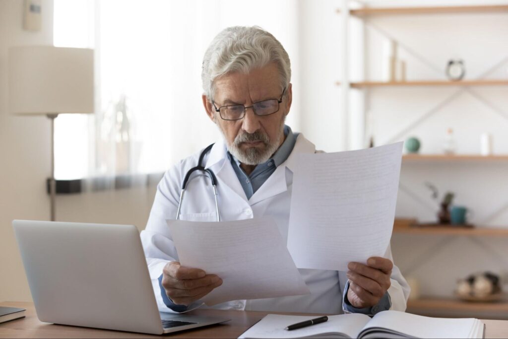 A white-haired, bearded physician sits at a desk with an open laptop as he reviews two documents.