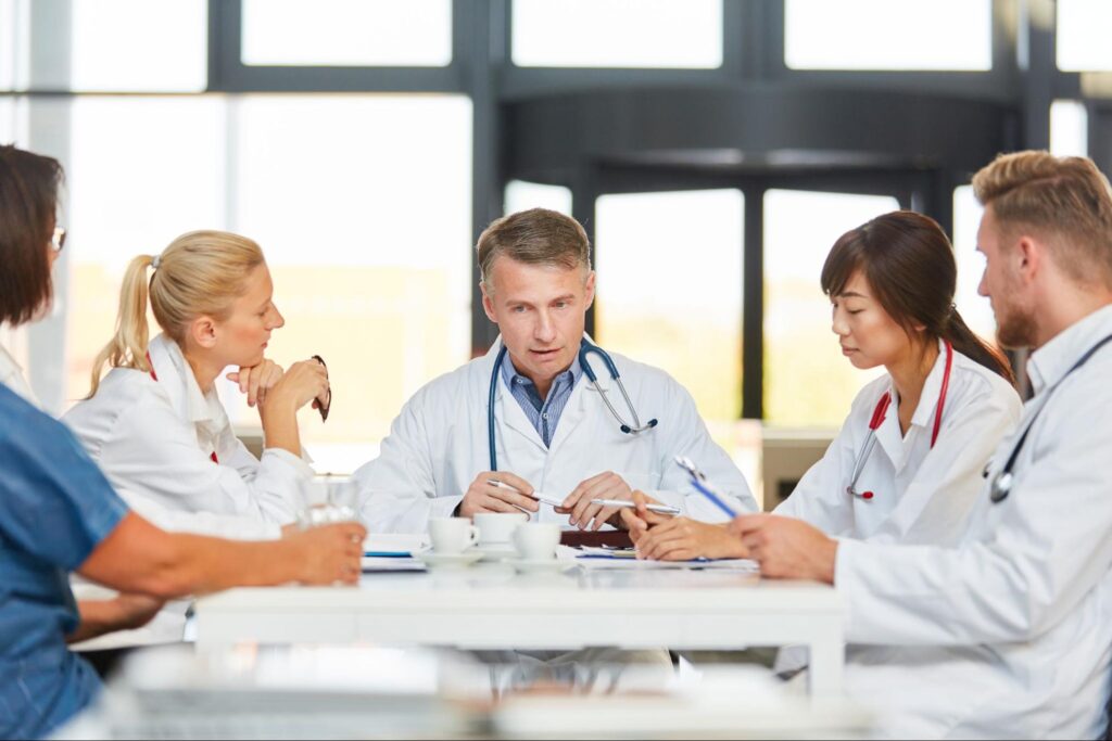 A group of five physicians sit at a table with coffee and clipboards.
