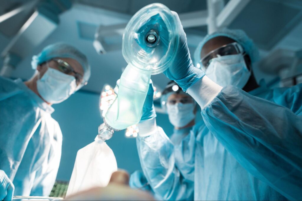 A CRNA lowers a mask onto a patient’s face while two other medical professionals stand by.