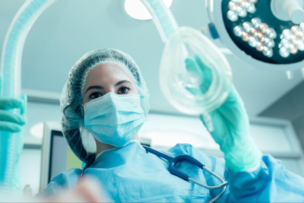 An anesthesiologist wearing blue scrubs lowers a mask toward the camera where her patient presumably lies.