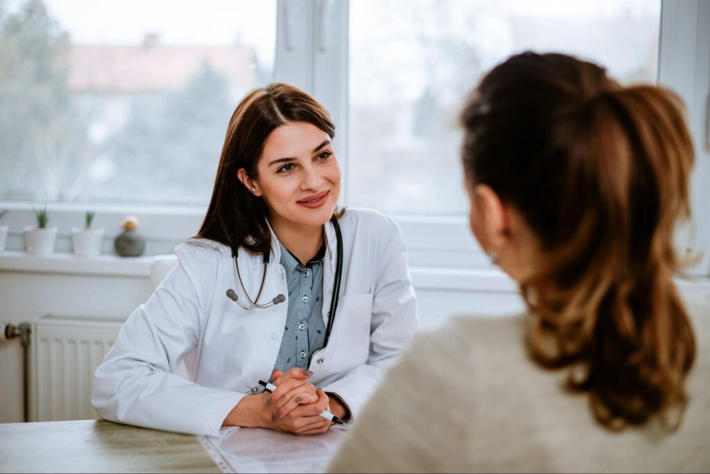 A woman in a white coat is facing the camera where a woman in a beige shirt sits across from her during an onsite physician interview.