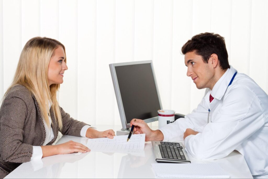 A blonde woman in a blazer sits across a desk from a physician in a white coat during an interview.