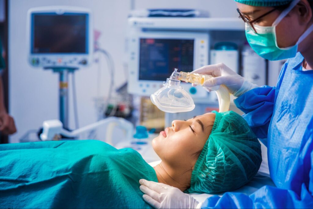A patient lies on an operating table while an anesthesiologist lowers a breathing mask onto their face.
