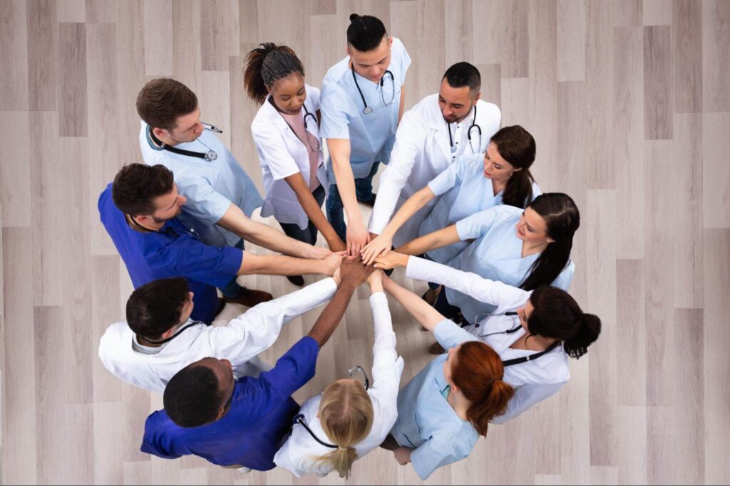 A group of twelve diverse healthcare professionals stand in a circle, placing their hands together at the center.