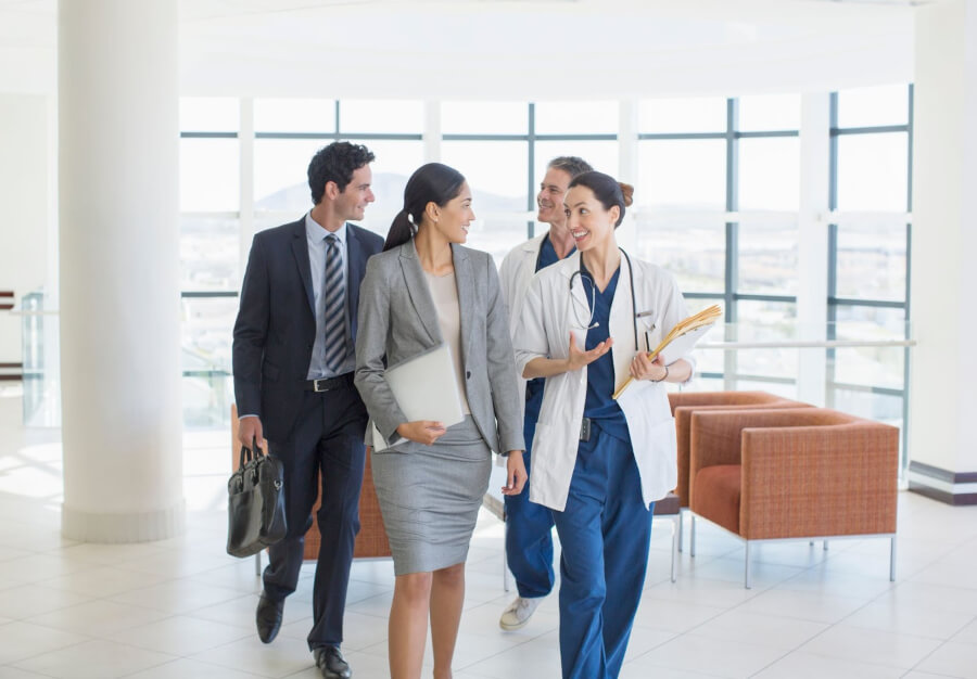 Two medical professionals guide two individuals in professional apparel through a medical facility as part of their candidate visit to hire a doctor.