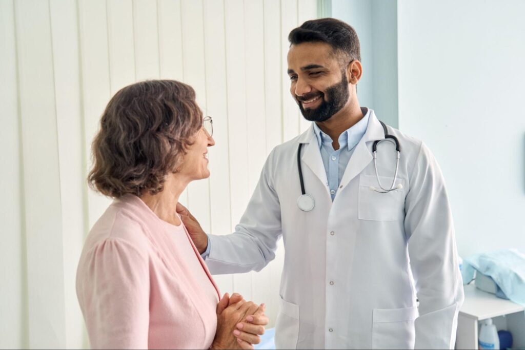 A male physician engages with an older patient. They are both smiling as he rests a hand on her shoulder.