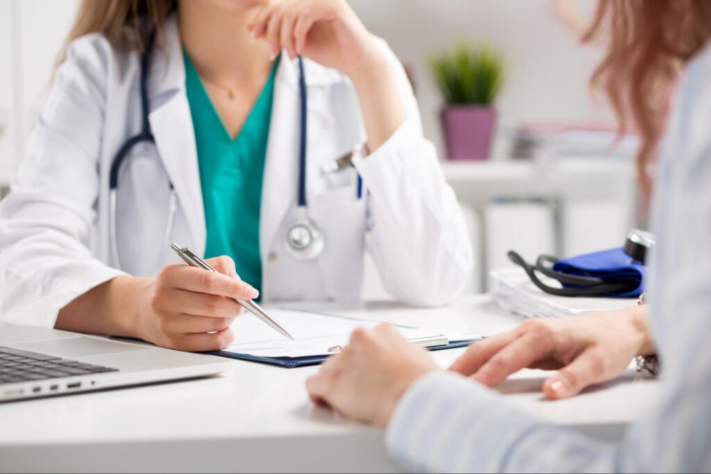 A woman in a white physician’s coat over scrubs speaks with a patient.